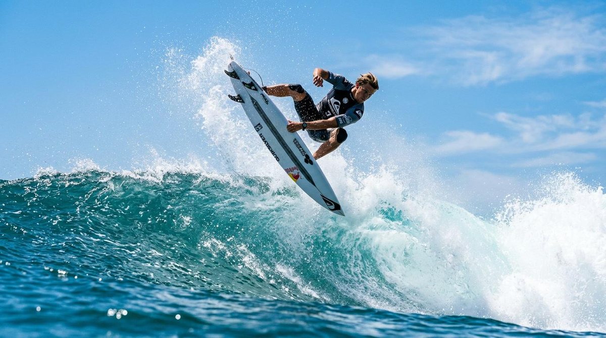 Ein Surfer im Sprung über dem Kamm einer Welle, Gischt, Meer und Himmel in strahlendem Blau.