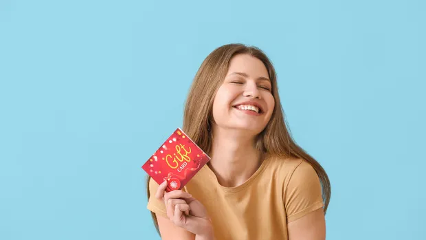 A smiling young woman holds a red gift card up beside her face, against a light blue background.