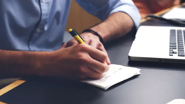A man in a blue shirt jots down information on a notepad, next to a laptop.