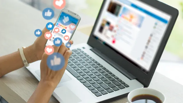 A person scrolls through a news feed on a smartphone surrounded by “like” icons, in front of a laptop open on a social network.