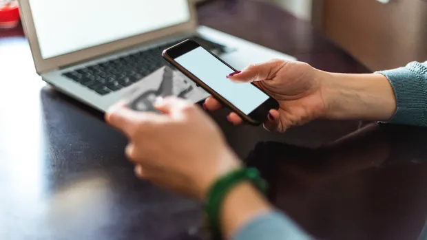 A person holds a smartphone in one hand and a ticket in the other, in front of an open laptop on a table.