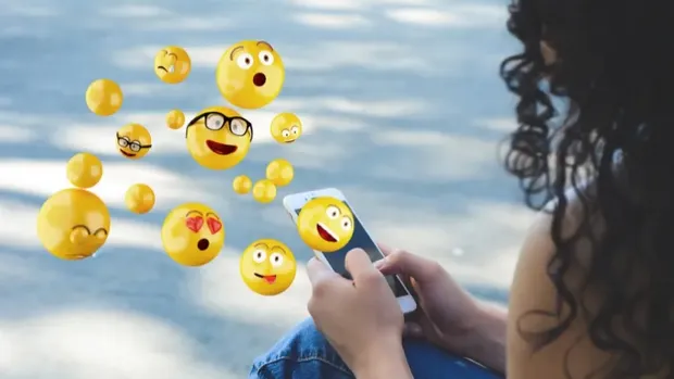A young woman looks at her smartphone, from which smiling emojis fly out.