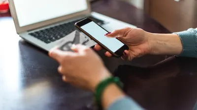 Eine Person hält ein Smartphone und ein Ticket vor einem Laptop, der auf einem Tisch steht.