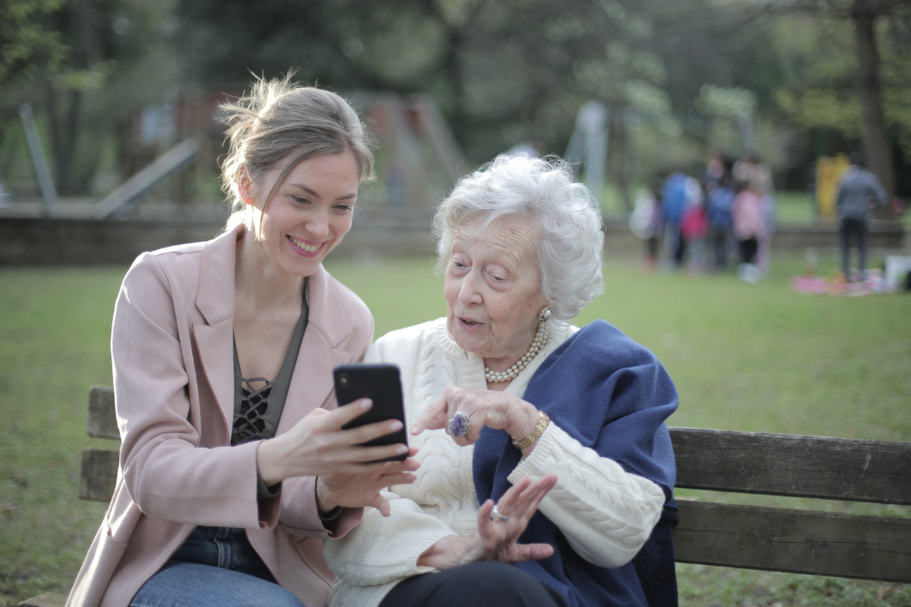Lächelnde junge Frau, die einer älteren Dame neben ihr auf einer Parkbank ein Smartphone zeigt