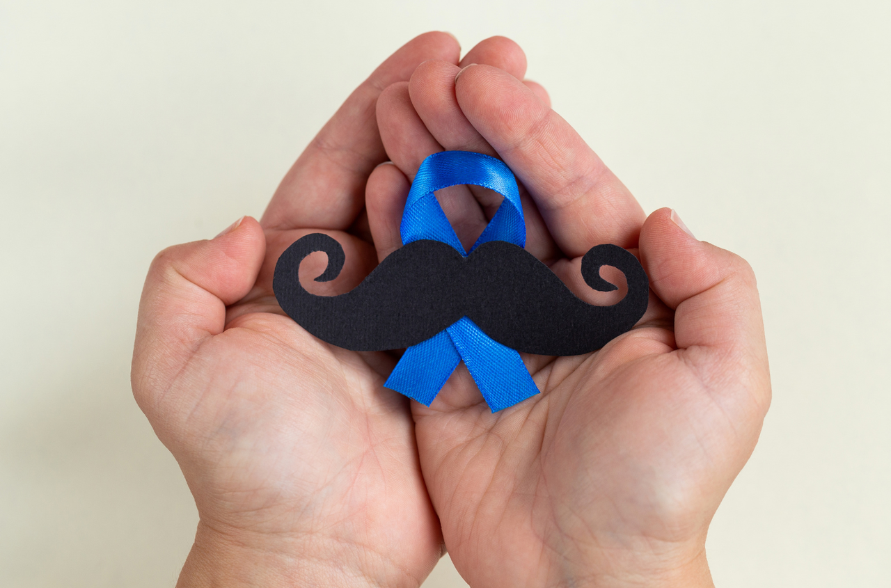 Dwie otwarte dłonie trzymające czarne papierowe wąsy i niebieską wstążkę Movember