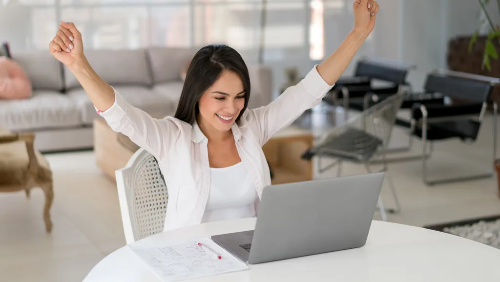 A woman sitting in front of her laptop raises her arms in a sign of victory.